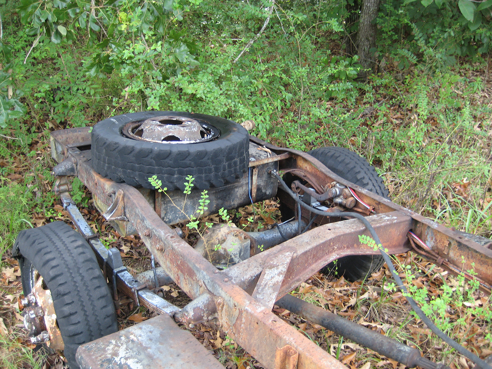Back half of rusted chassis of 1941 Army Command Car with an extra tire, surrounded by overgrown weeds and grass.