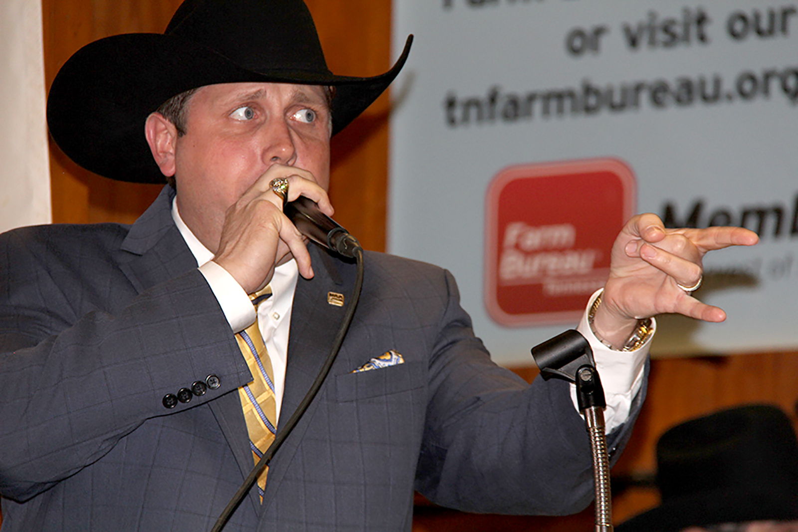 Auctioneering man in a grey suit and black cowboy hat speaking into a handheld microphone and pointing to the right of the frame.