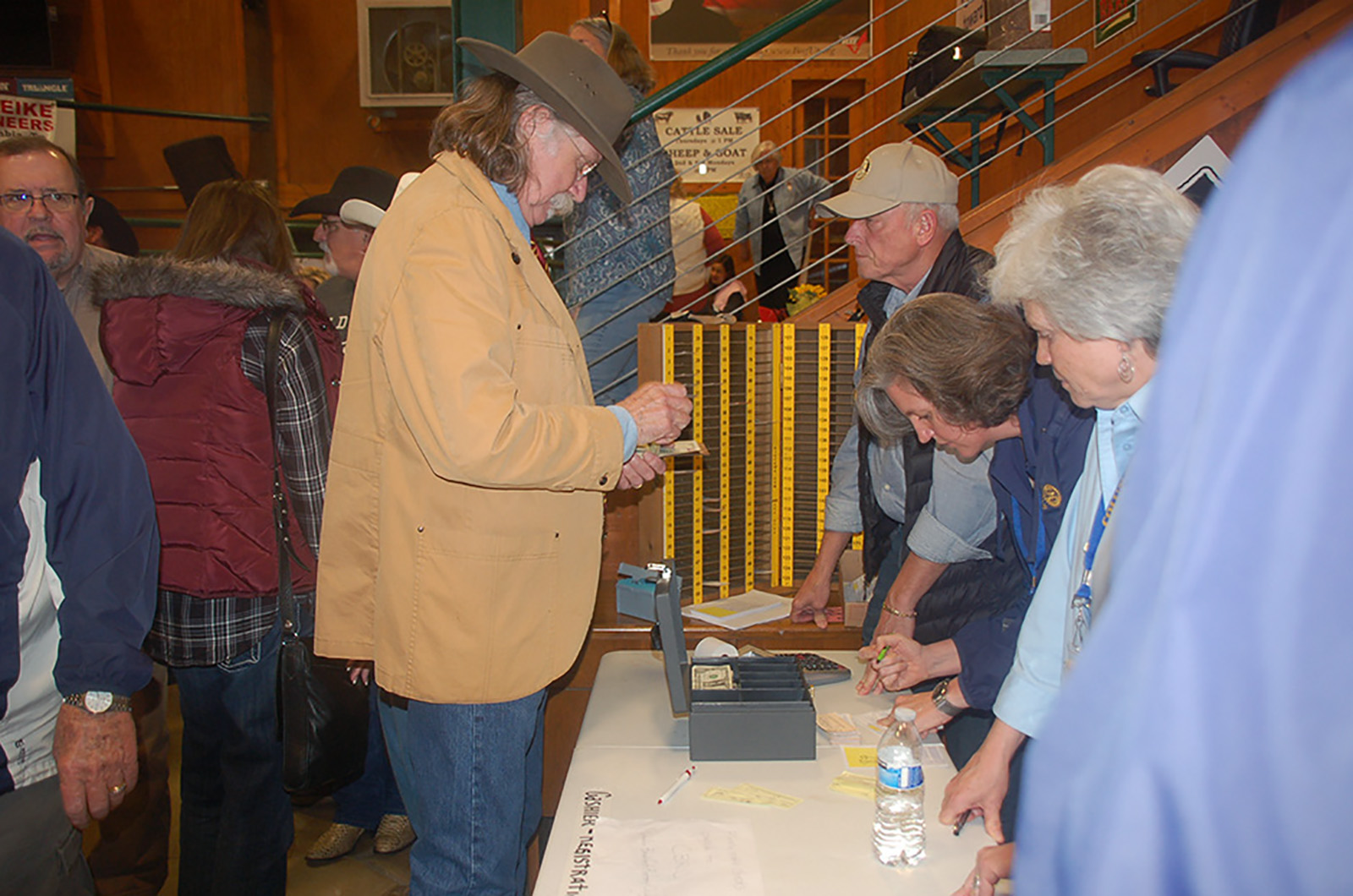 Colonel Littleton, wearing a tan jacket, blue jeans, and hat, is writing a check at the accounting table at an auction.