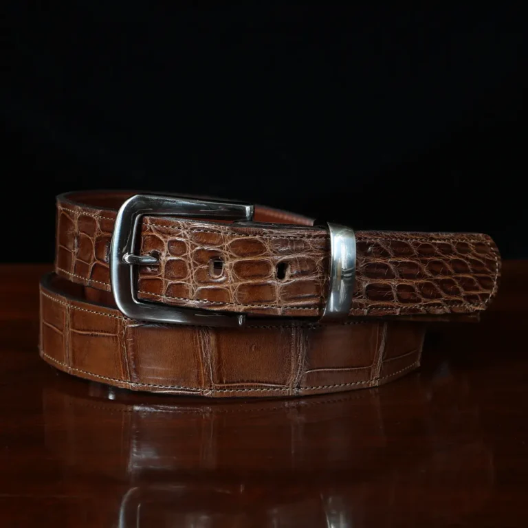 An American Alligator Belt with Nickel hardware showing the front coiled view on a wooden table with black background