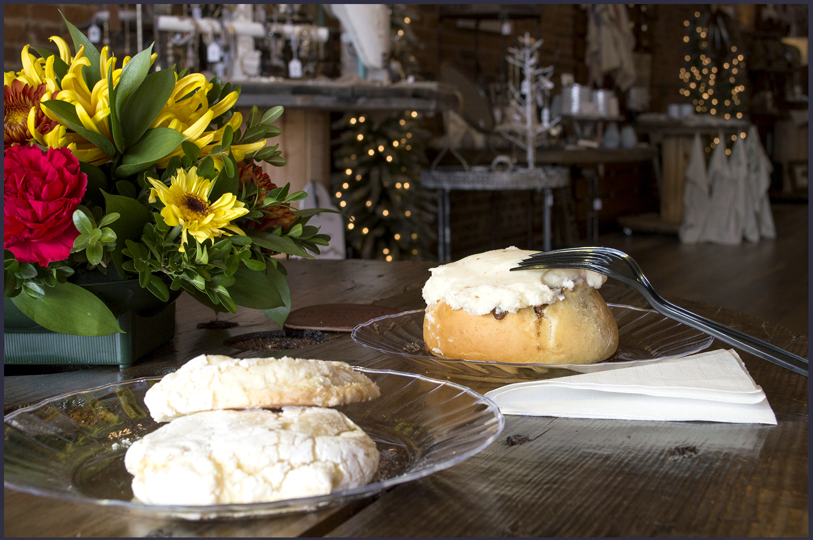 Two lemon butter cookies on a clear plastic plate in the front left and a large icing-covered cinnamon roll on a plate behind it to the right with a pot of flowers to the far left.