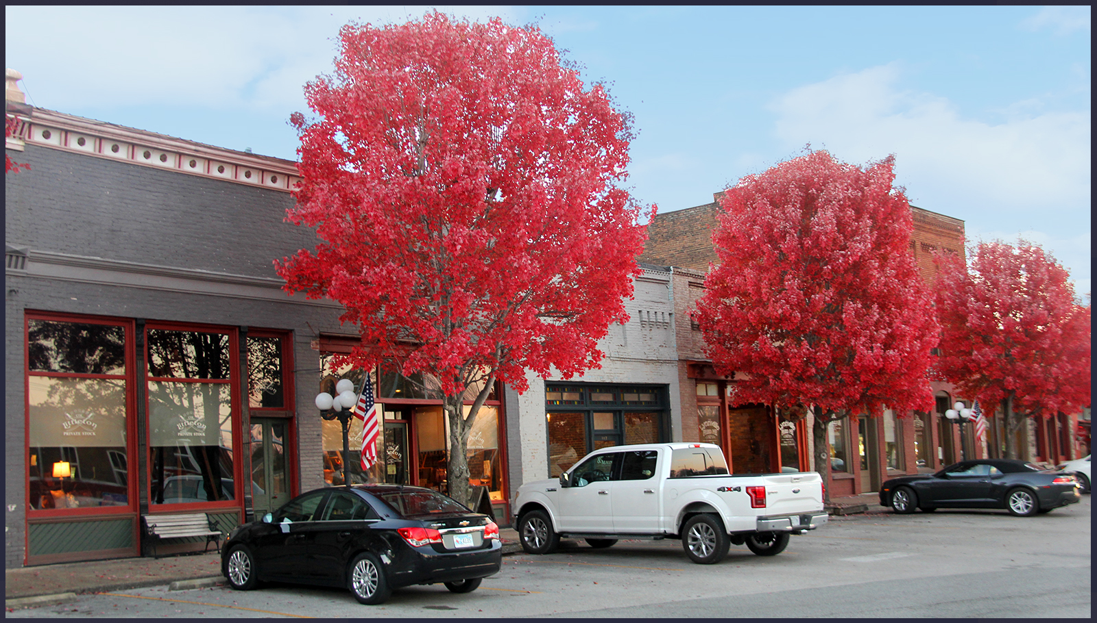 The downtown Lynnville town row, a few vehicles parked out front, and the three maple trees in view are full and red-leafed