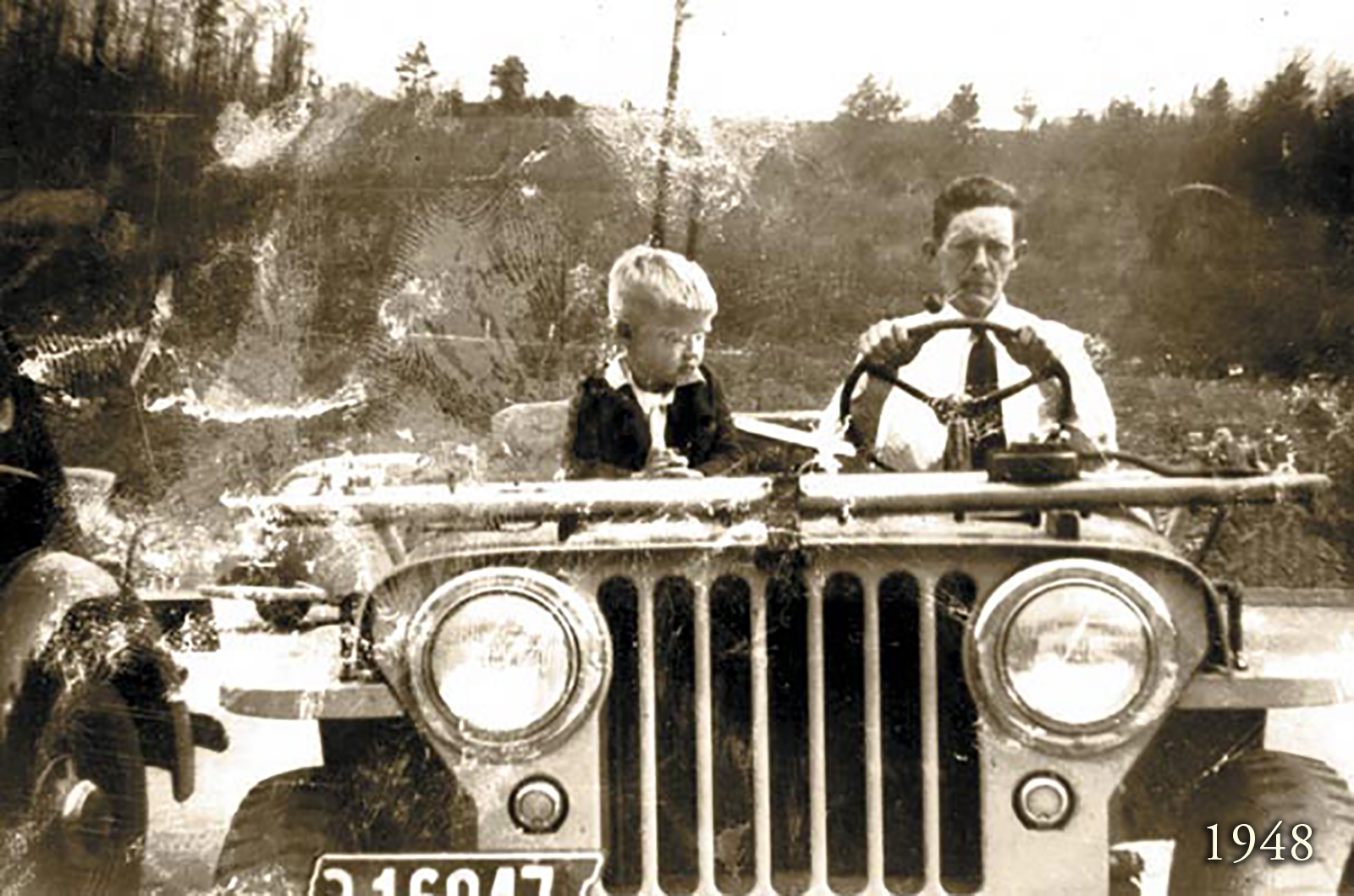 Sepia toned image of a young Colonel Littleton riding in an old jeep with his dad in 1948