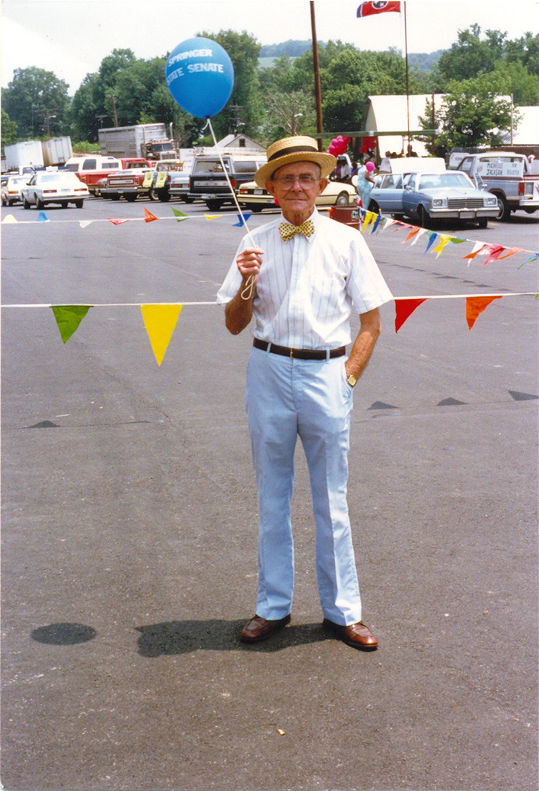 Colonel's dad standing on asphalt holding a blue balloon and wearing a bowtie and carnival barker's hat