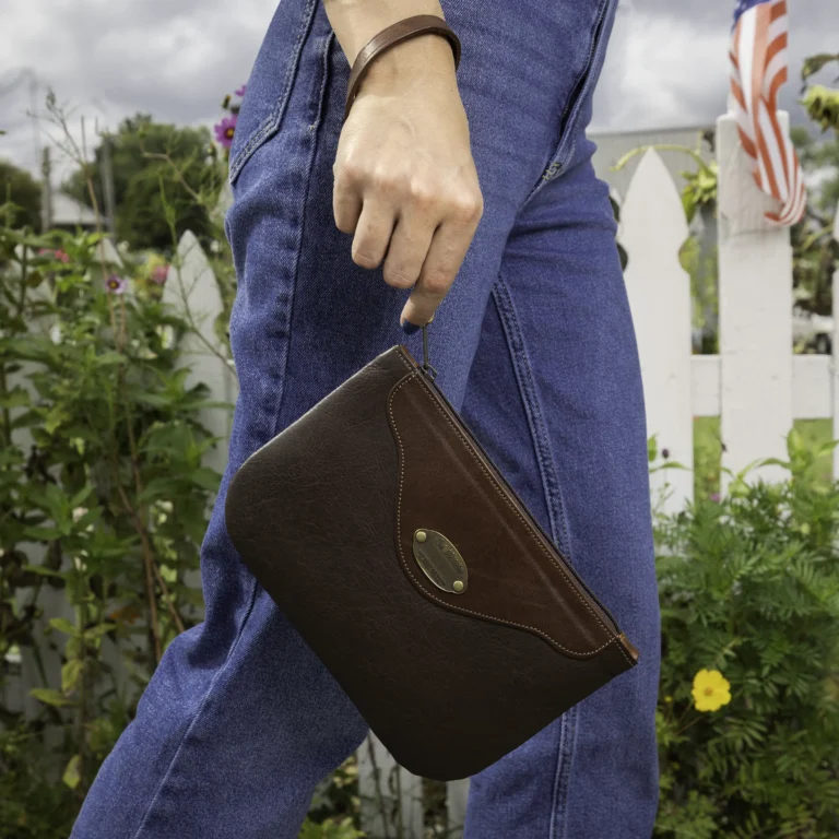 woman carrying a brown leather wristlet clutch in front of a fence