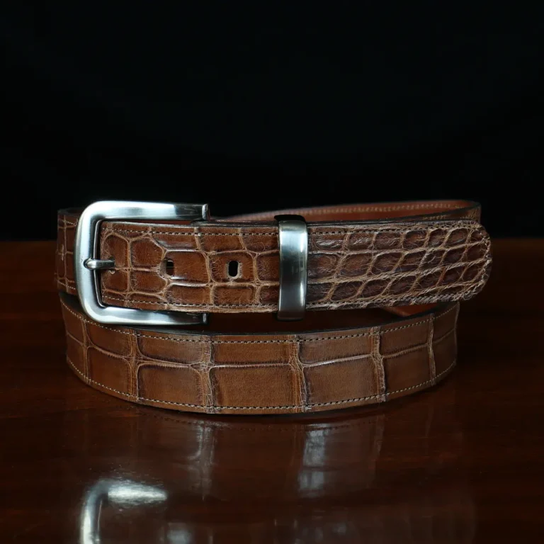 An American Alligator Belt with Nickel hardware showing the front coiled view on a wooden table with black background